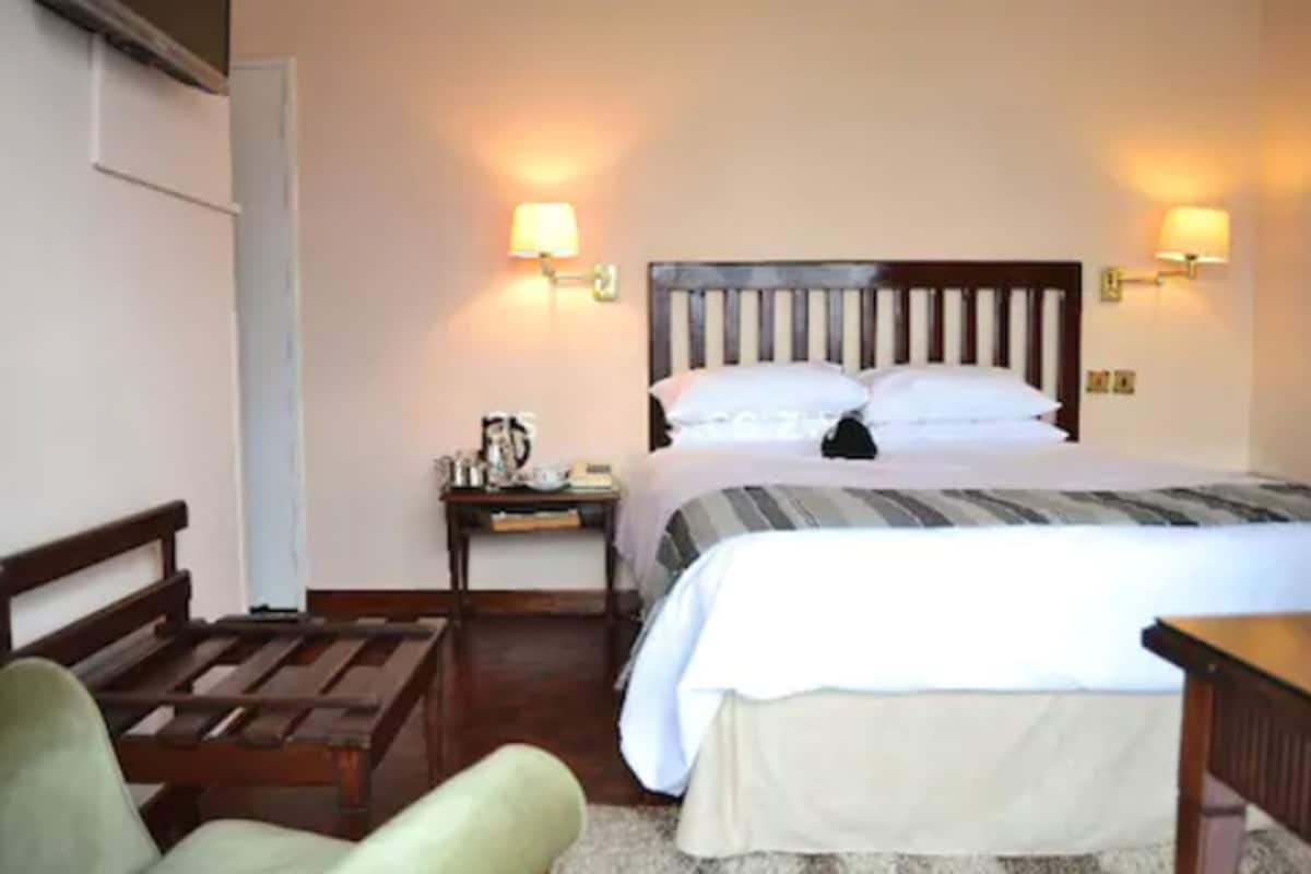A hotel room with a wooden bed, two bedside lamps, a nightstand with a lamp, a green armchair, and a wooden table.
