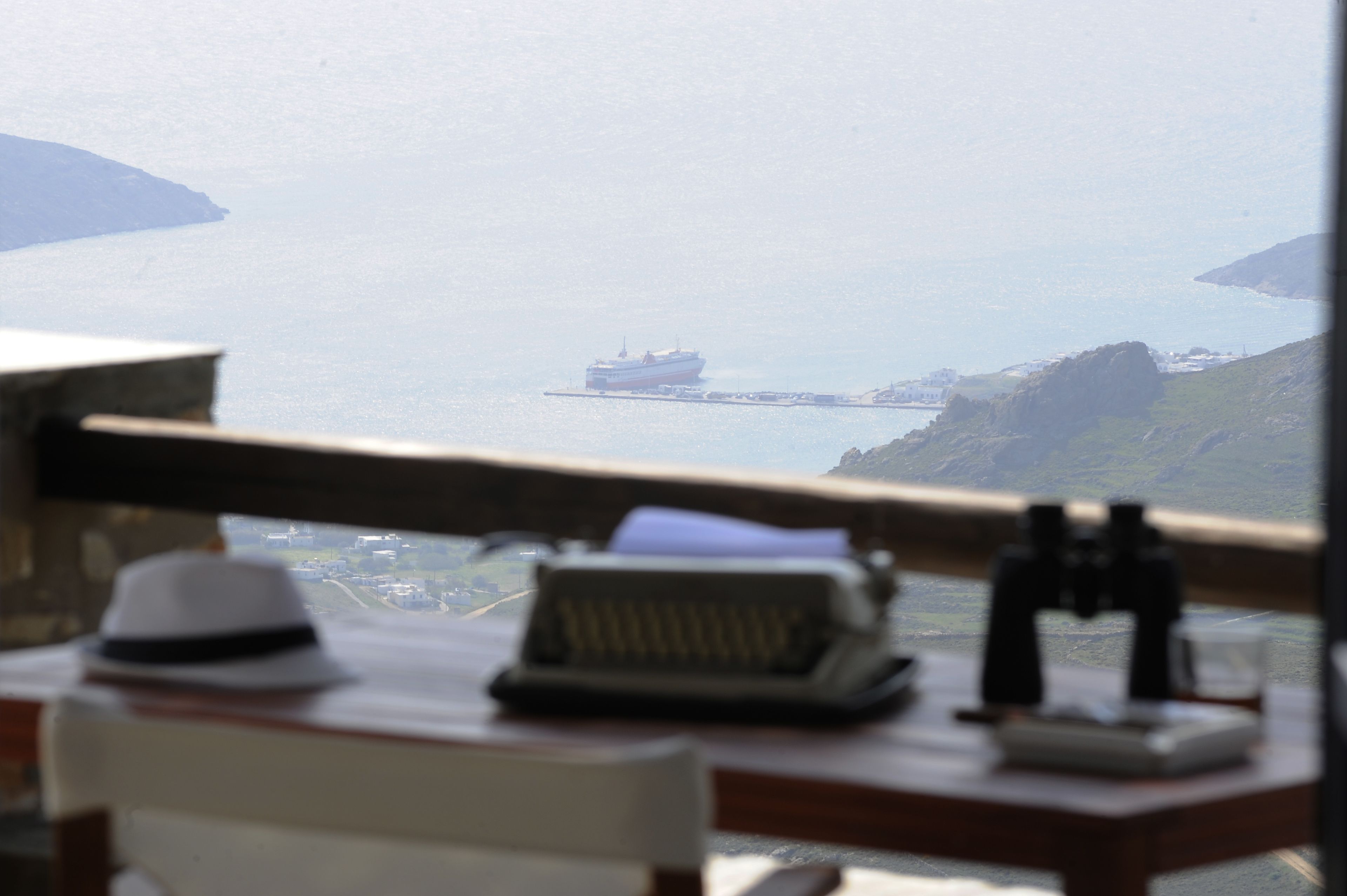 Un bureau avec une machine à écrire, un chapeau et des jumelles, donnant sur une vue côtière avec un navire et une terre lointaine.