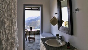A bathroom with a stone wall, a white sink, a mirror, and a view of mountains through a doorway. - Aria Villas (Serifos)