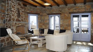A stone-walled room with a white sofa, a wooden chair, and a table with a book and candle. - Aria Villas (Serifos)