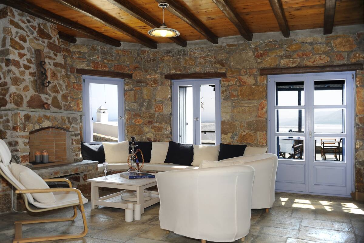 A stone-walled room with a white sofa, a wooden chair, and a table with a book and candle.