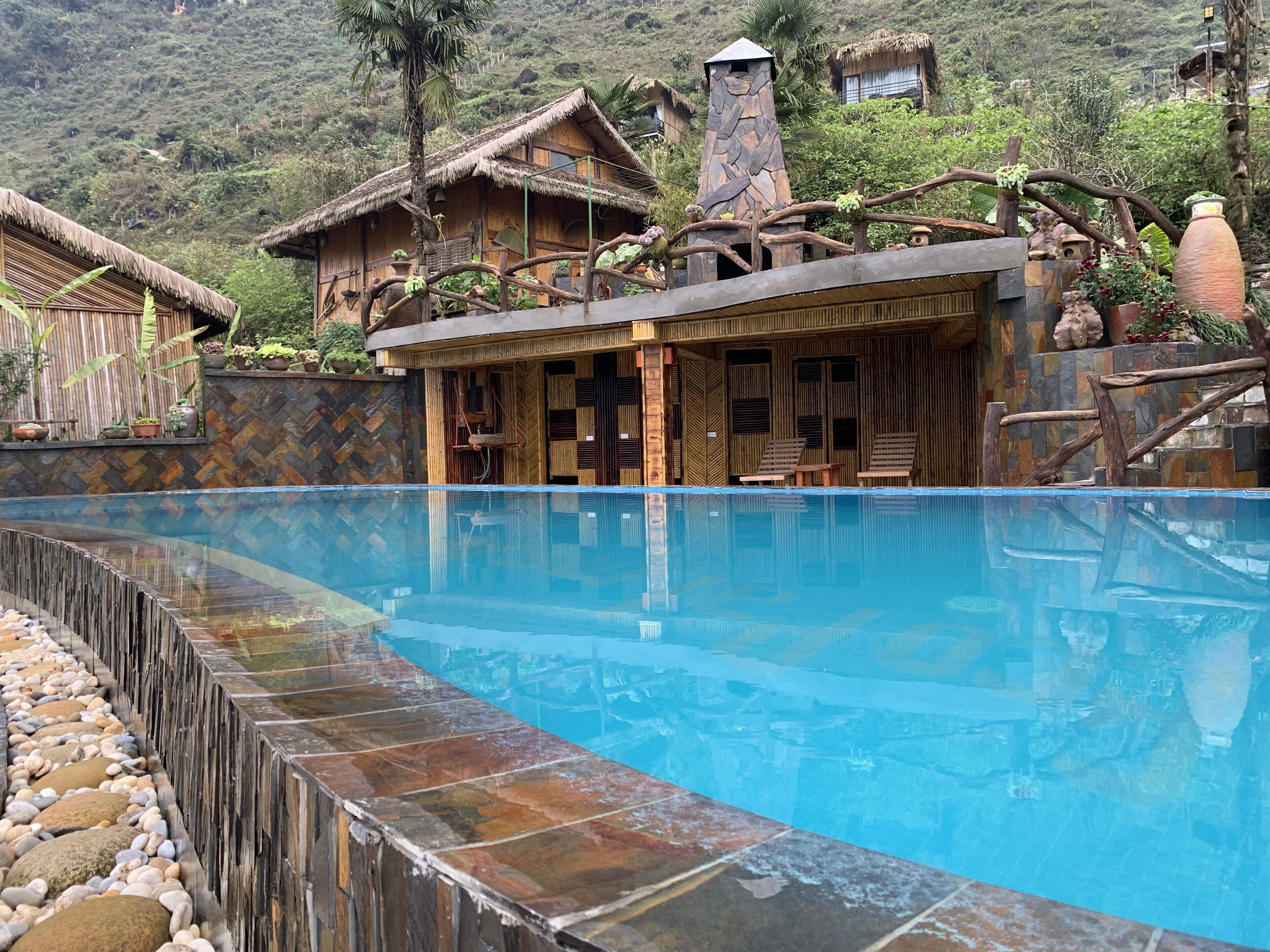 A pool area with a stone border, wooden deck, and thatched-roof buildings in the background.