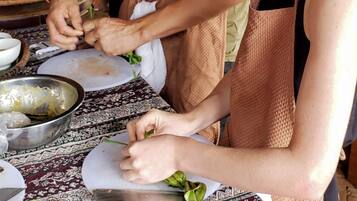 Zwei Personen bereiten Essen an einem Holztisch mit gemustertem Tischläufer zu, umgeben von Küchenutensilien und Zutaten.