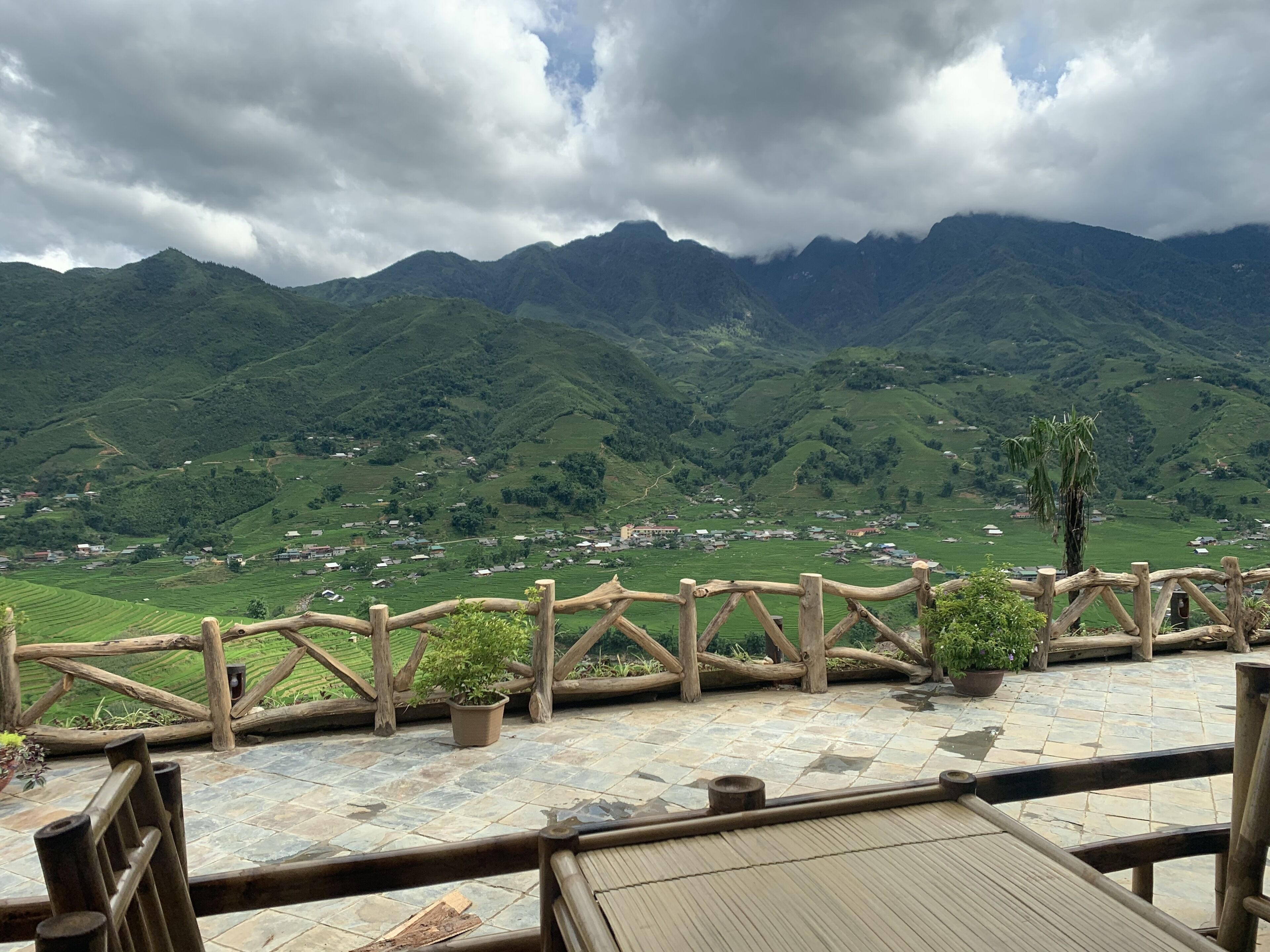 A terrace with a wooden railing, potted plants, and a view of green hills and a village.