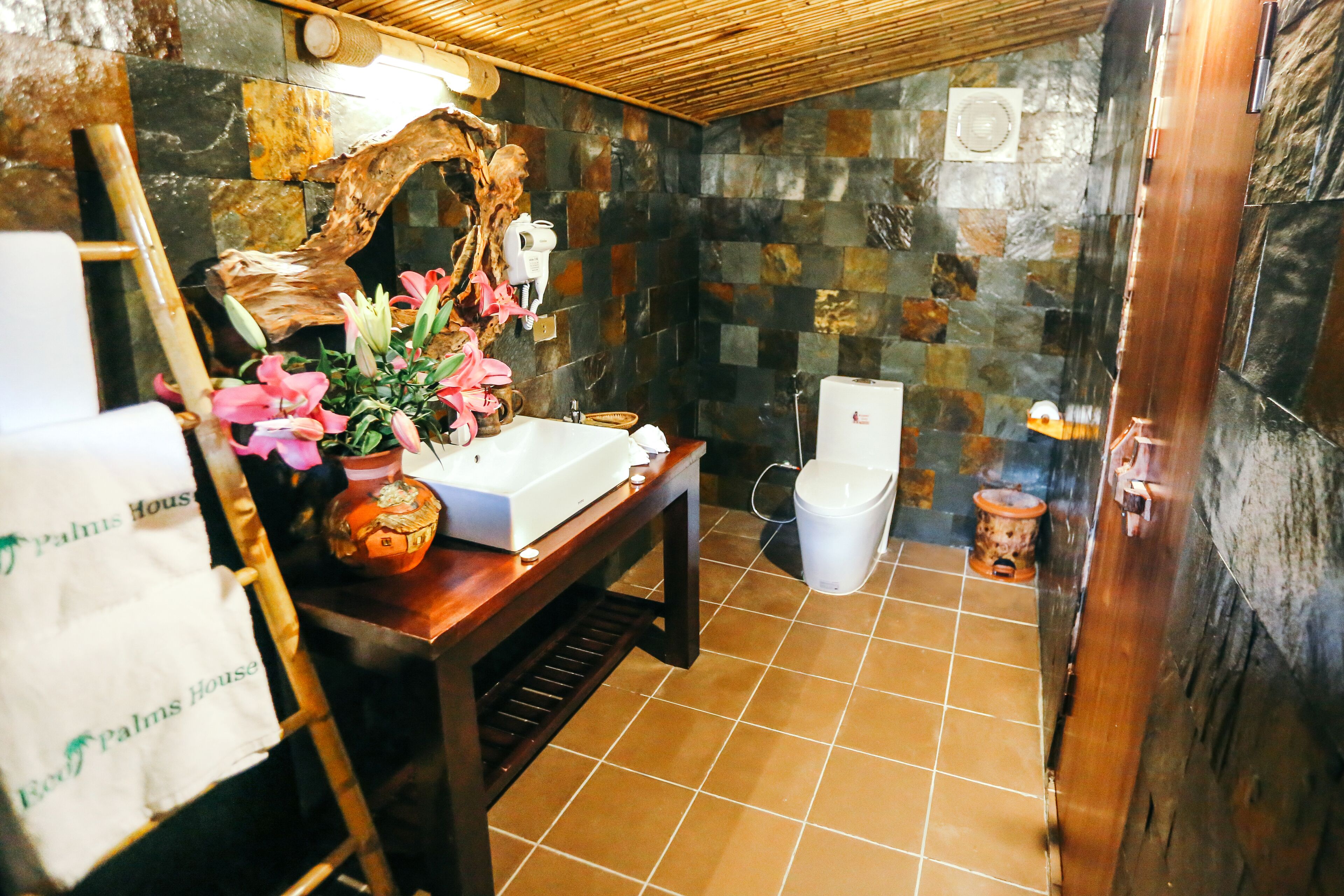 Een badkamer met een stenen muur, een houten bankje, een wastafel met een bloevas en een toilet.