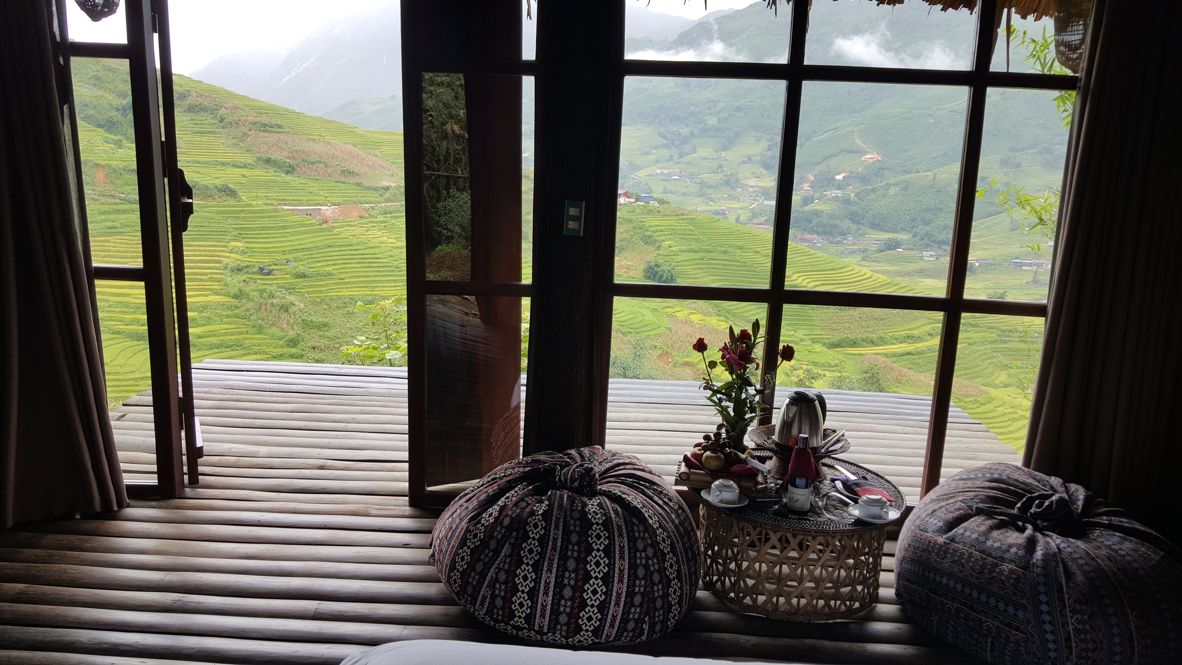 Quarto com grande janela dando para campos em socalcos. Há piso de madeira, almofada estampada e pequena mesa redonda com um vaso de flores.