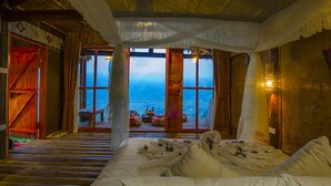 Een kamer met een bed, een bankje en uitzicht op zee. Er zijn grote ramen en een houten plafond.