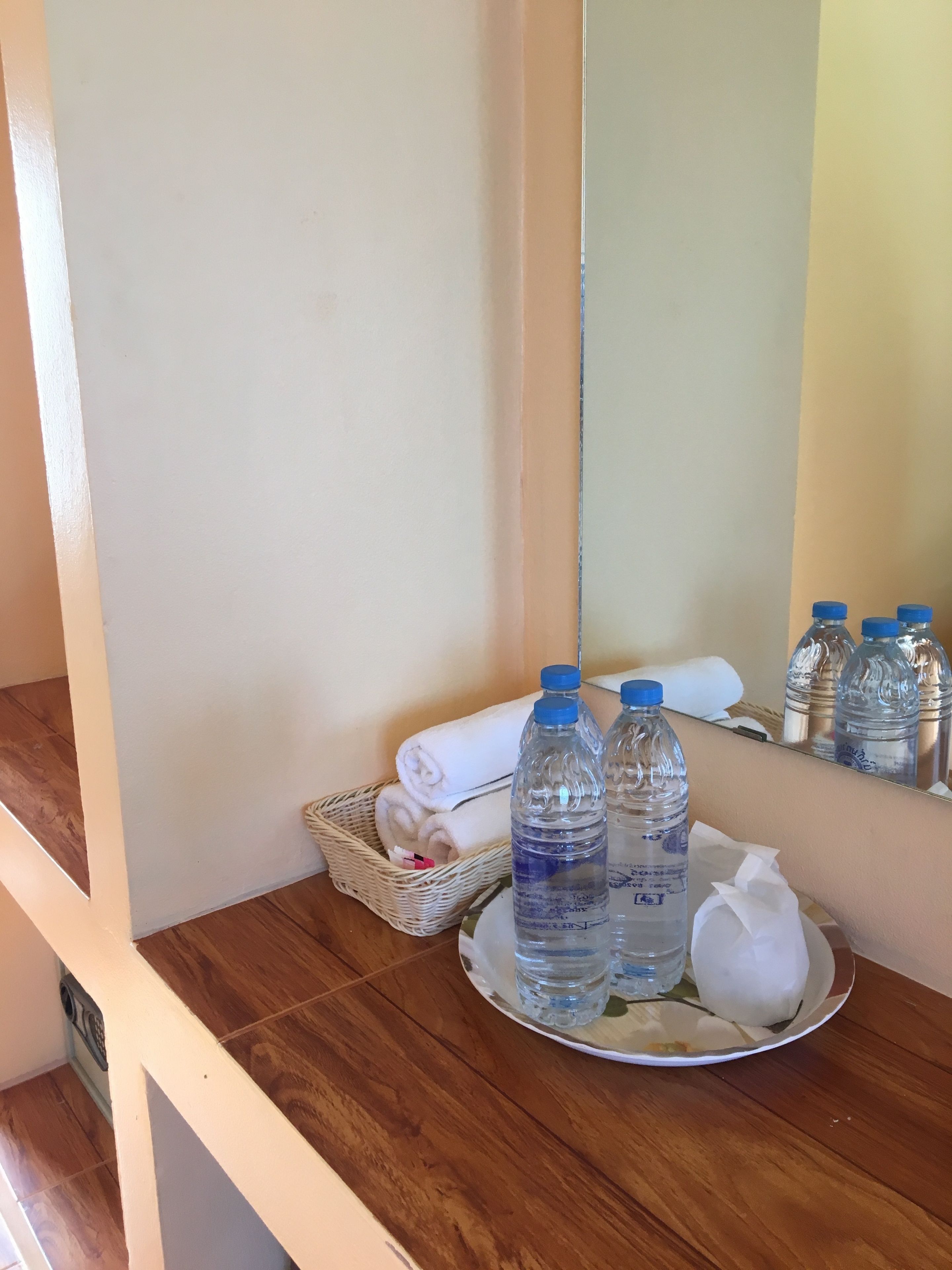 Un baño con piso de madera, una toalla blanca, una cesta de mimbre y tres botellas de agua sobre un plato.