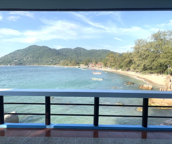 A coastal view from a balcony with a railing, clear blue sea, sandy beach, and forested hills in the background.