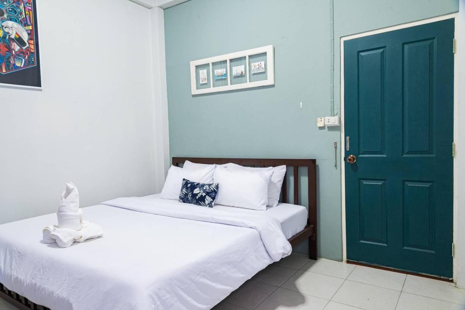 A neatly made bed with white linens and a folded towel, a wooden headboard, a teal-colored door, and a wall with framed pictures.