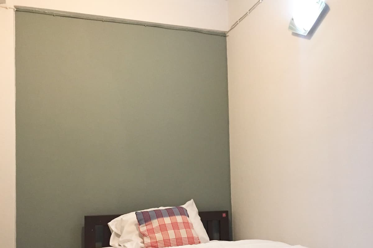 Una cama bien hecha con ropa blanca y una almohada de cuadros, un interruptor de luz en la pared y una pared lisa con un detalle en color verde.