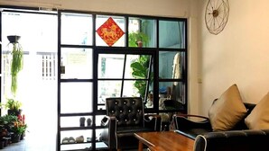 A modern living room with a black sofa, wooden coffee table, and a black leather chair. There is a large window with a decorative red sign, and a wall clock. - Asoke Montri Hostel (Bangkok)