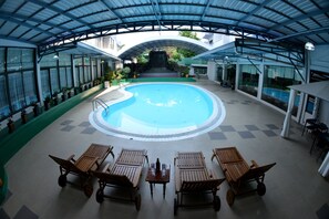 A covered swimming pool with lounge chairs and tables, surrounded by potted plants and a waterfall view. - Golden Butterfly Hotel (Yangon)