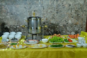 A buffet table with various dishes, a large silver pot, and a tiled wall in the background. - Golden Butterfly Hotel (Yangon)