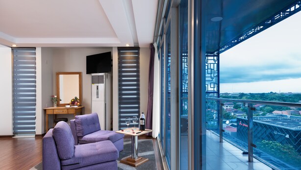 A modern living room with a purple sofa, a small table with wine glasses, a TV, and a city view through large windows. - The Hub Hotel (Yangon)