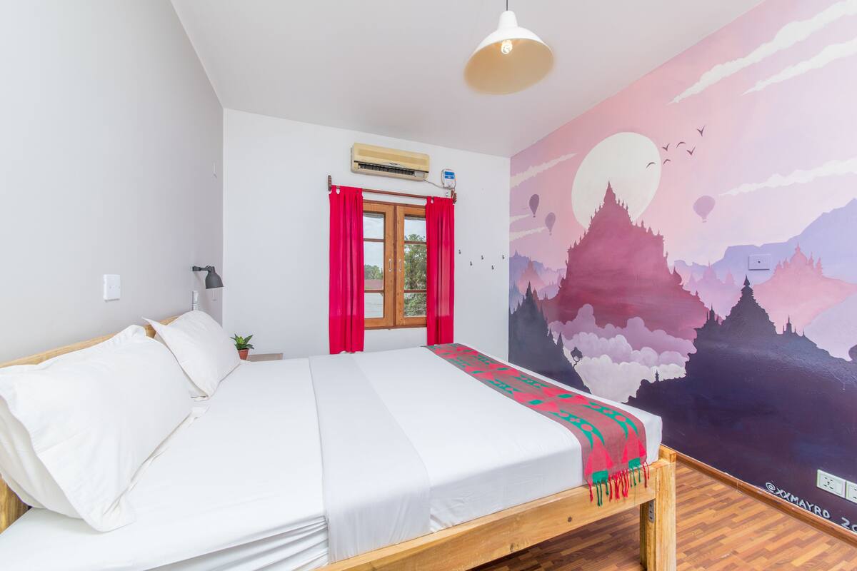 Ein Schlafzimmer mit einem Bett, einem Fenster mit roten Vorhängen, einer Klimaanlage und einem Wandgemälde.