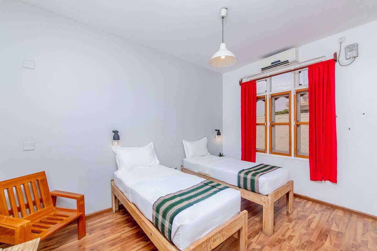 Ein Zimmer mit zwei Holzbett, einer Holzbank, einer weißen Wand, einem roten Vorhang und einer Deckenleuchte.