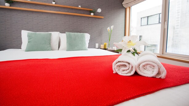 Ein ordentlich bezogenes Bett mit einer roten Decke, zwei grünen Kissen und einem Strauß weißer Blumen.