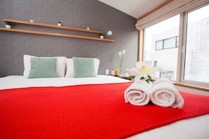 Uma cama bem arrumada com um cobertor vermelho, duas almofadas verdes e um arranjo de flores brancas.