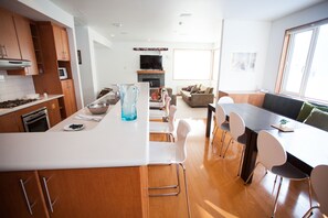 A modern kitchen with white countertops, wooden cabinets, and a central island. There is a dining area with a table and chairs, and a living room with a sofa and a fireplace. - Shirokuma 1 (Kutchan)