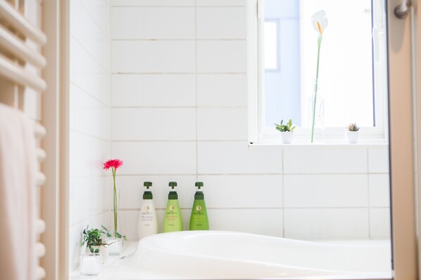 A bathroom with a white bathtub, a window with a view, and three green bottles on the wall.