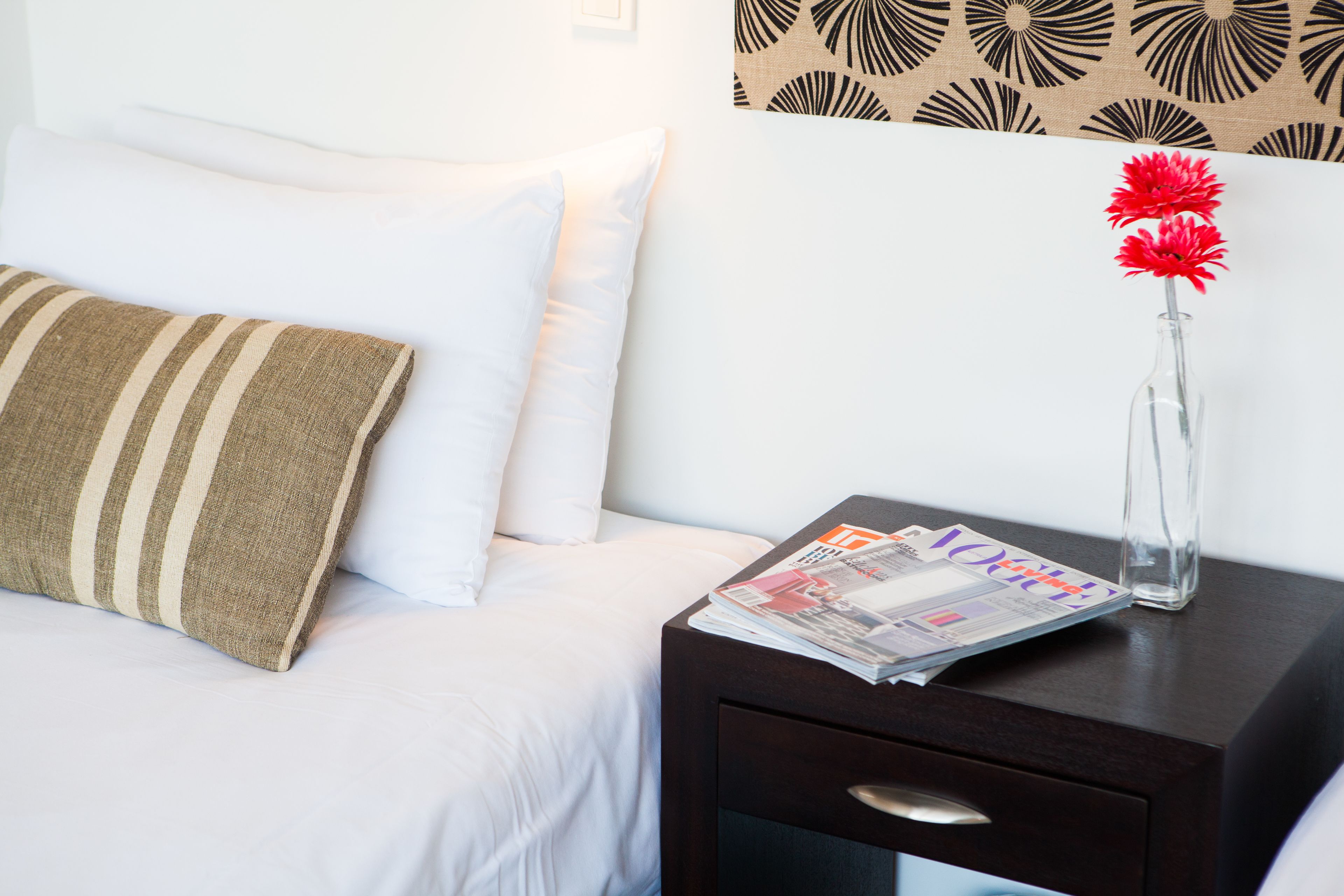 Un lit bien fait, avec des oreillers blancs et rayés, une table de chevet sombre sur laquelle repose un magazine, et un vase rempli de fleurs rouges.
