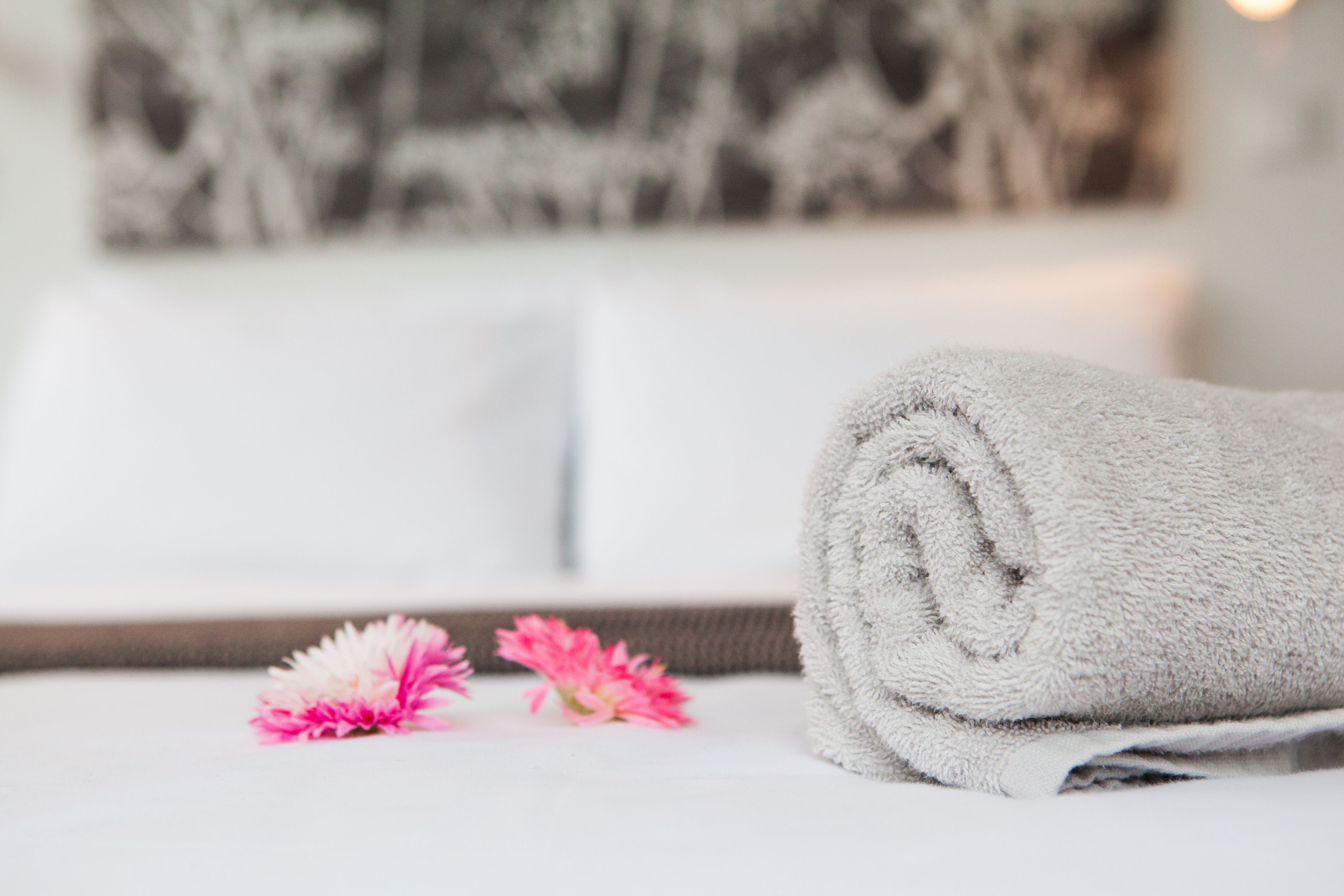 A neatly folded white towel on a bed with a white pillow and a wall art piece in the background.