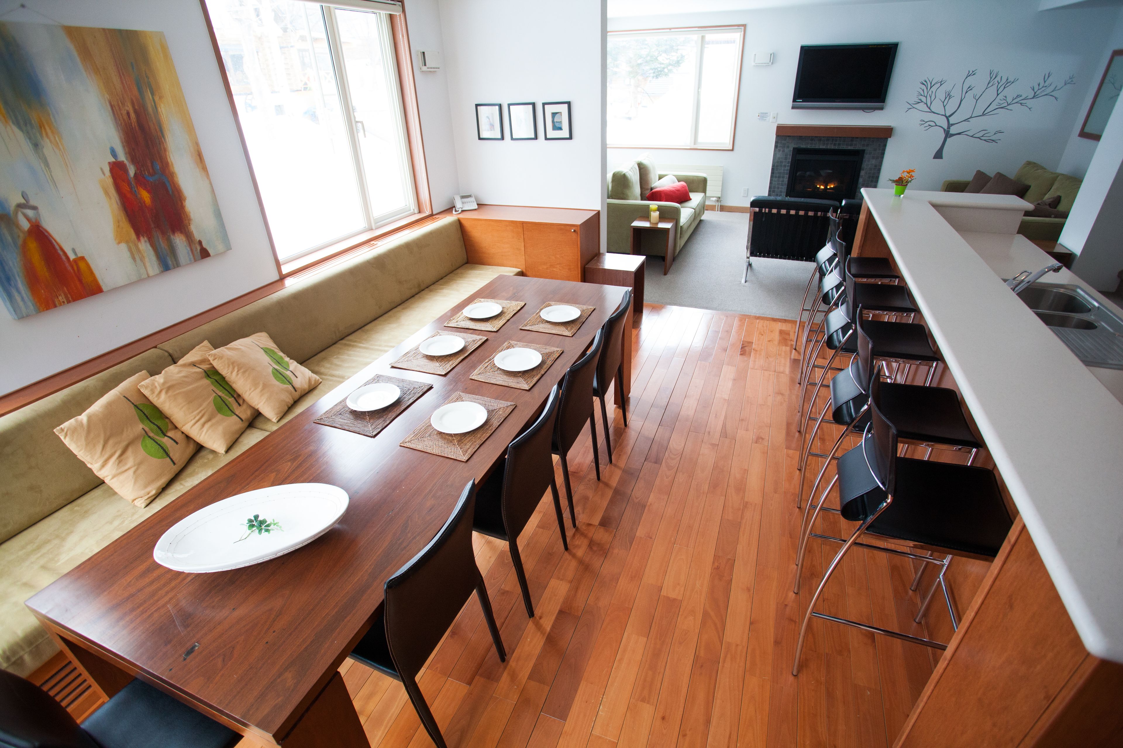 Una sala de estar moderna con una mesa de comedor de madera preparada para cuatro personas, un sofá con cojines, una chimenea, un televisor montado en la pared y un amplio ventanal que proporciona luz natural.