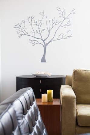 Una habitación con un sofá de cuero negro, un sillón color beige, una mesita de madera con velas y un adhesivo de pared con diseño de árbol.