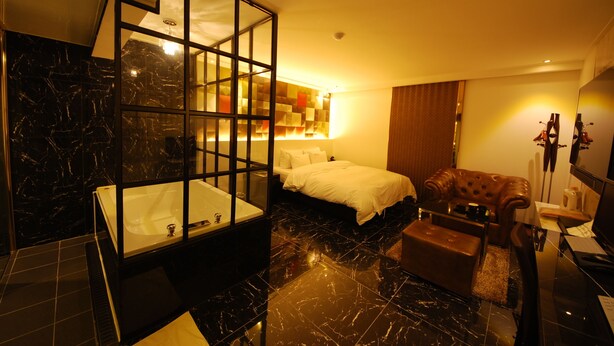 A modern bathroom with a large bathtub, a separate shower area, and a glass-enclosed shower. There is a bed with white bedding and a brown leather armchair in the room. - Hotel BUTI & (Incheon)