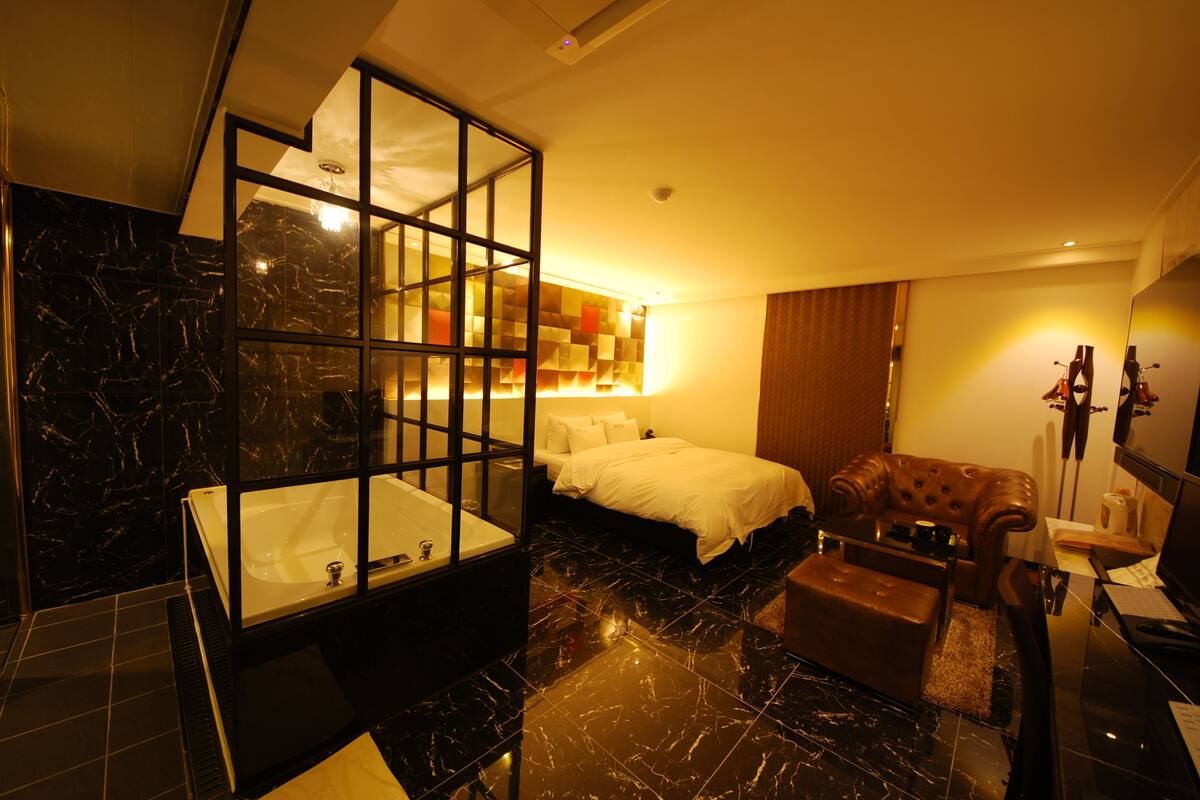 Une salle de bain moderne avec une grande baignoire, un espace douche séparé et une douche à l’italienne avec des parois en verre. La chambre comprend un lit avec une literie blanche et un fauteuil en cuir marron.