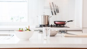 Un plan de travail de cuisine avec un saladier blanc contenant des fruits, un verre d’eau et une planche à découper avec un couteau.