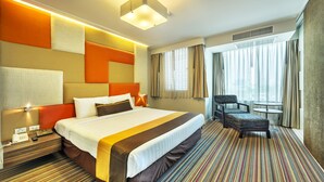 Quarto de hotel com uma cama grande, uma mesinha de cabeceira com telefone, uma cadeira, uma mesa pequena e uma janela com cortinas.