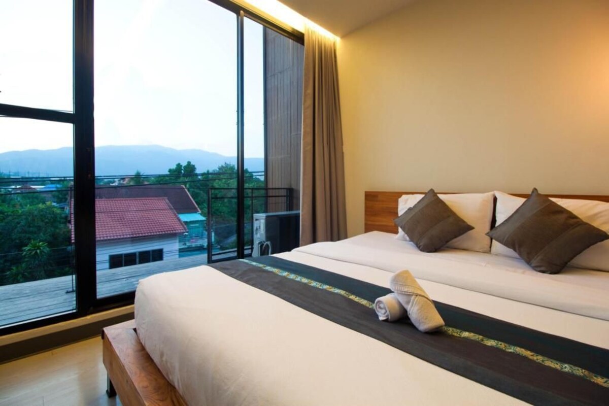 A hotel room with a large window, a bed with pillows, a bench, and a view of a building and mountains.