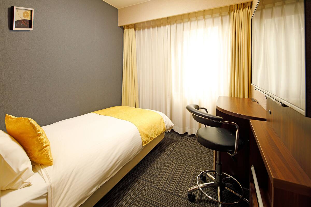 A hotel room with a single bed, a desk with a chair, and a large window with curtains.