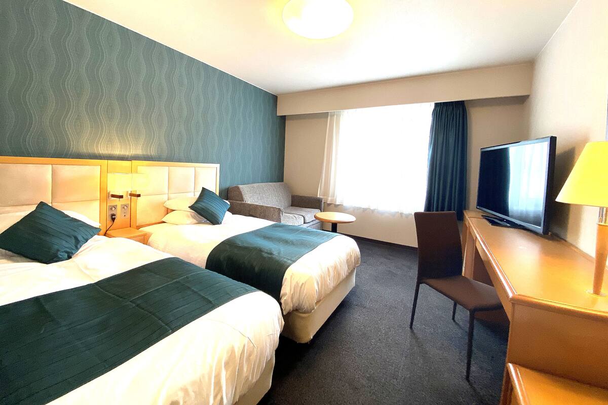 A hotel room with two beds, a desk with a TV, a chair, and a window with curtains.