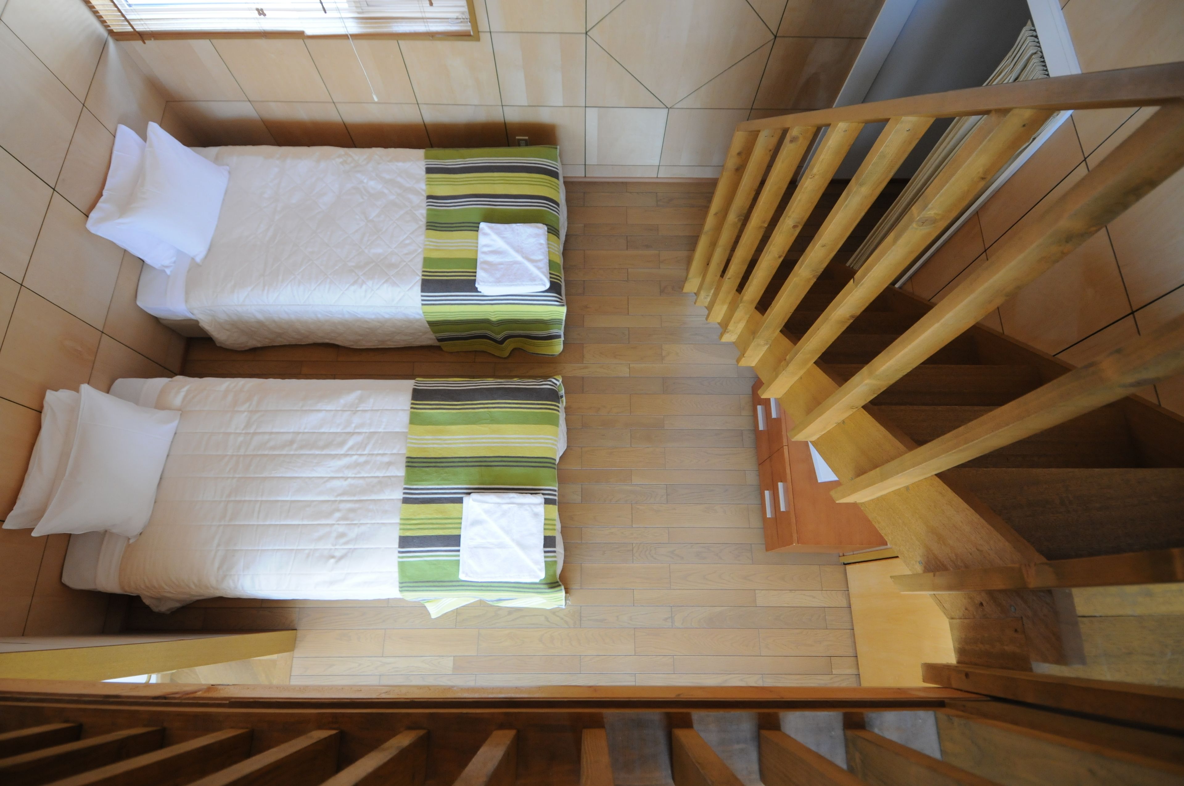 A room with two beds, wooden flooring, a staircase, and a window.