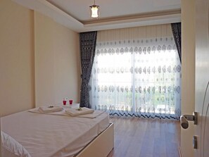 A hotel room with a bed, a bedside table, a mirror, and a window with patterned curtains.