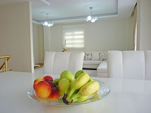 Een eettafel met een schaal fruit in een moderne woonkamer met witte meubels en een kroonluchter.