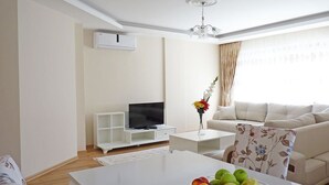 A modern living room with a white sofa, a flat-screen TV, a coffee table with a fruit bowl, and a chandelier. - White Star Antalya (Konyaalti)