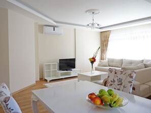 A modern living room with a white sofa, a flat-screen TV, a coffee table with a fruit bowl, and a chandelier. - White Star Antalya (Konyaalti)
