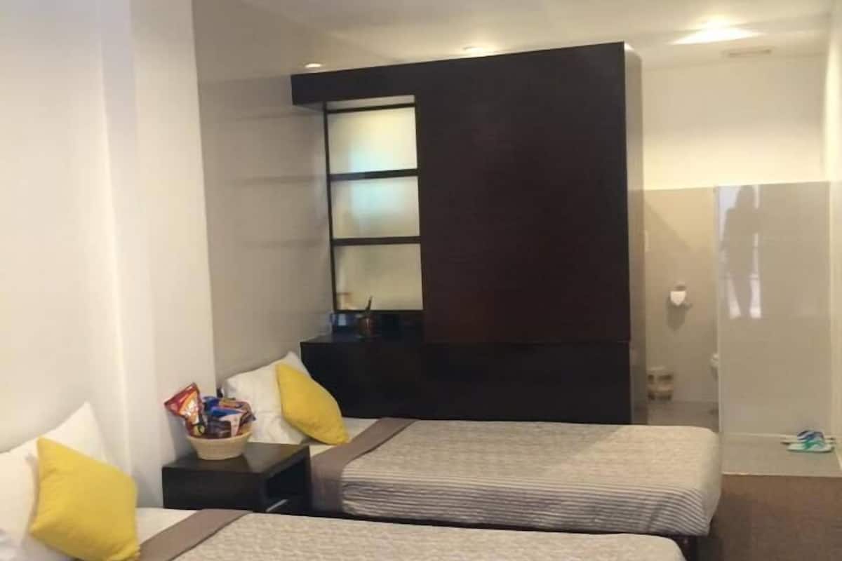 Una habitación de hotel con dos camas, una mesita de noche pequeña, una mesita auxiliar y un estante en la pared, además de una puerta.