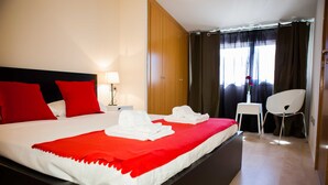 Una habitación de hotel con una cama grande, ropa de cama blanca y roja, un armario de madera, una pequeña mesa con una silla y una ventana con cortinas.