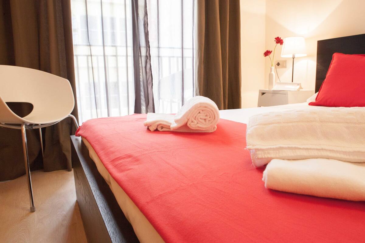 Ein Hotelzimmer mit Bett, Sessel, Nachttisch mit Lampe und einem Fenster mit Vorhängen.