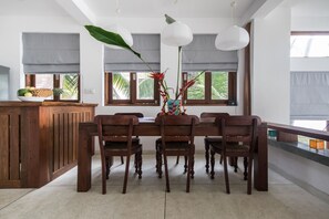 A dining area with a wooden table and chairs, a vase with flowers, and large windows with blinds. - Skinny Beach House (Unawatuna)