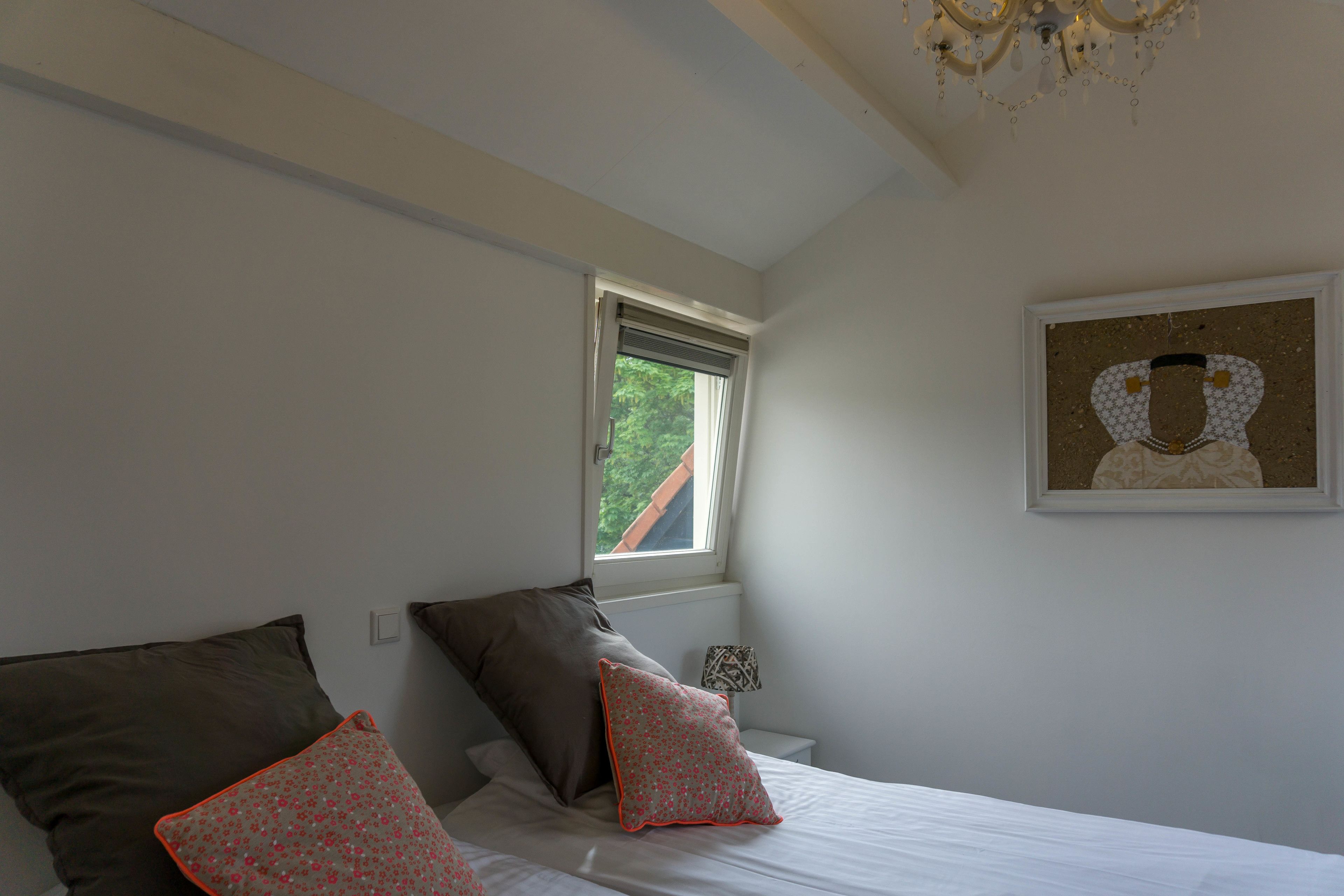 A bedroom with a bed, pillows, a nightstand, a lamp, a window, and a framed picture on the wall.