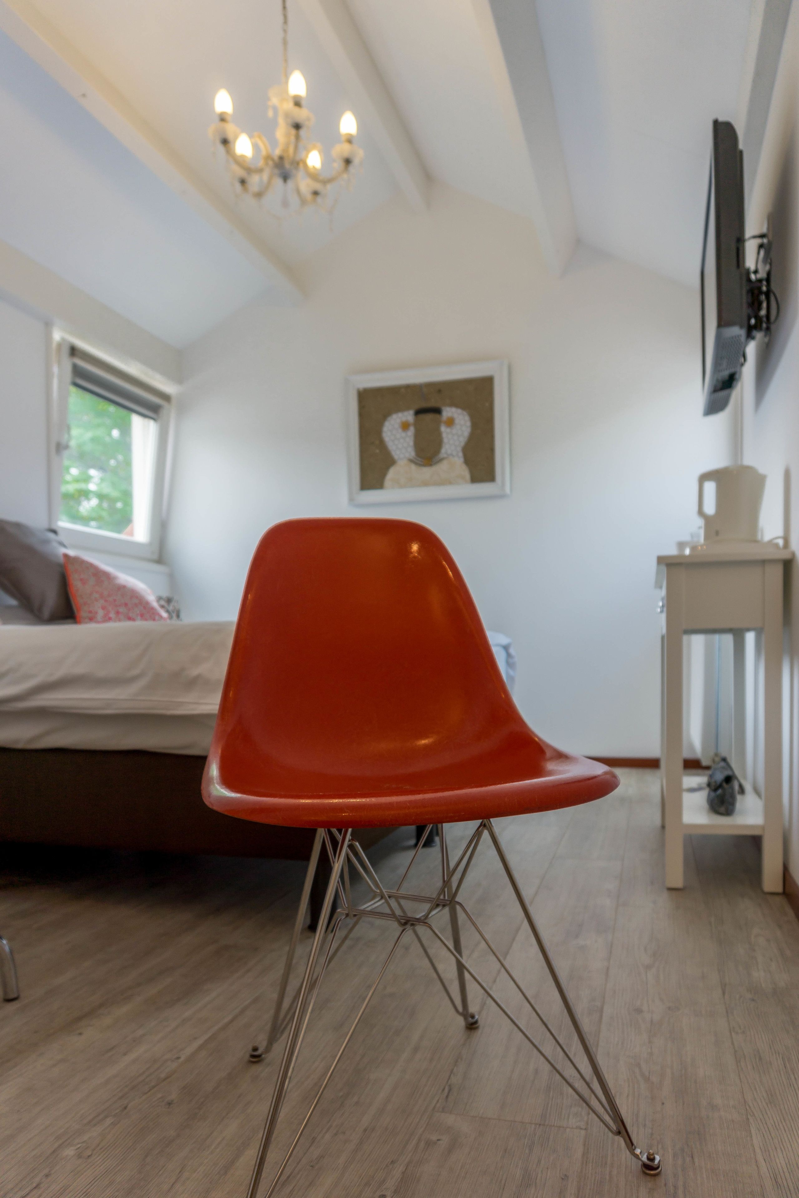 A bedroom with a bed, a red chair, a chandelier, a television, and a window.
