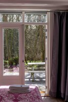 Una habitación con cama, ventana y puerta abierta que da a un espacio exterior con banco y árboles.