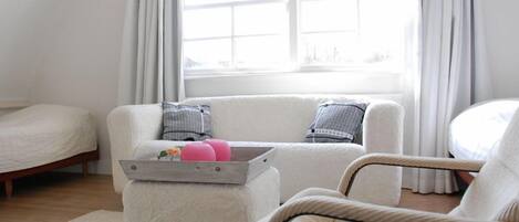 Une chambre avec un lit, un canapé et un fauteuil. Il y a une fenêtre avec des rideaux, et un plateau avec des objets roses sur le canapé.