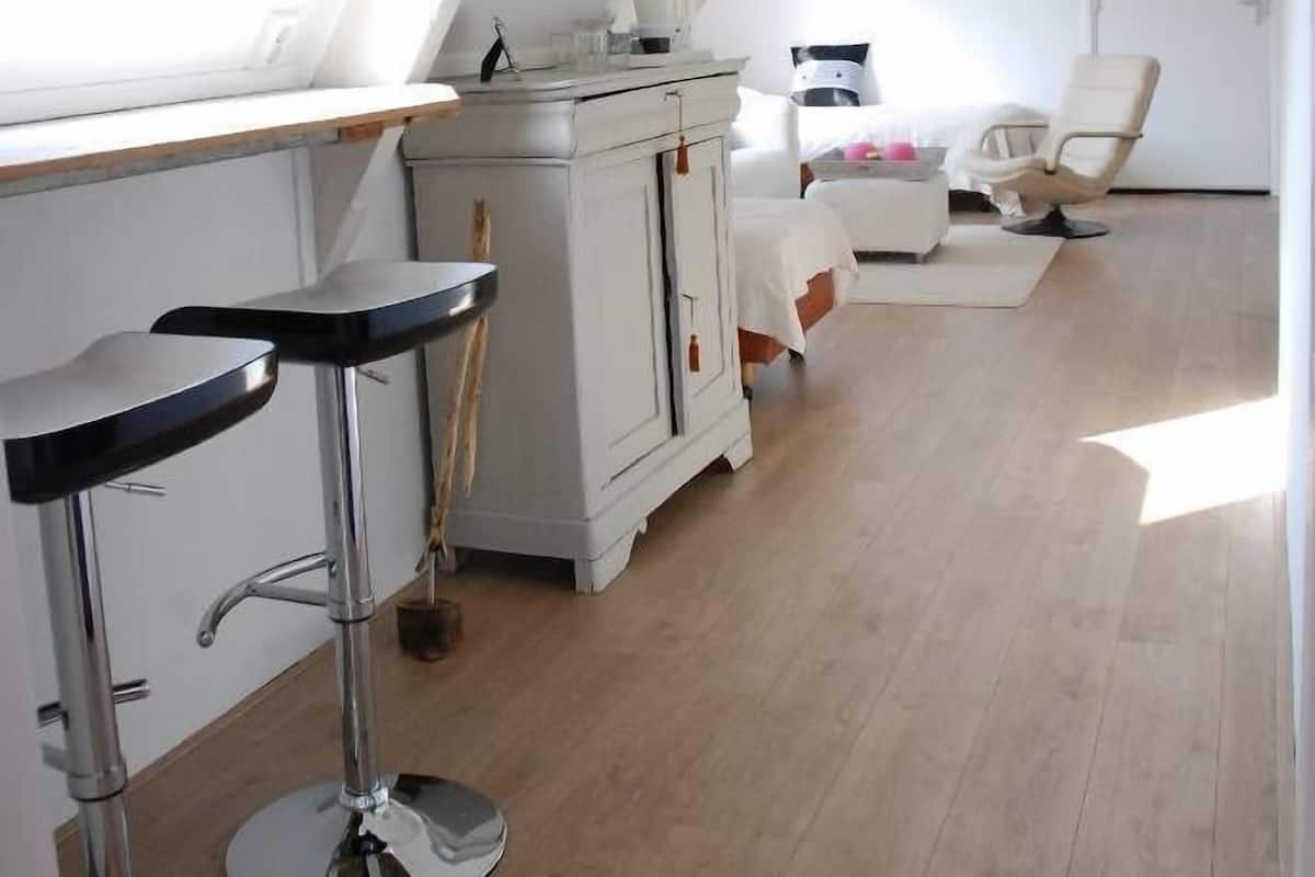 Een kamer met hoge plafonds, een dakraam, een wit bed, een massagestoel en een kleine kitchenette.